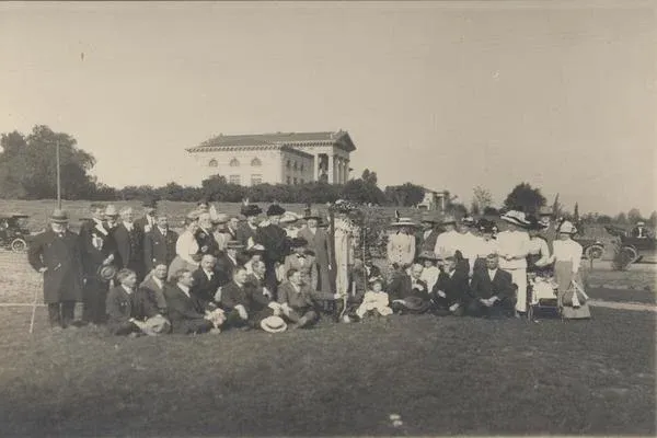 Group of people post with a large building in the background, sepia image, early 20th century